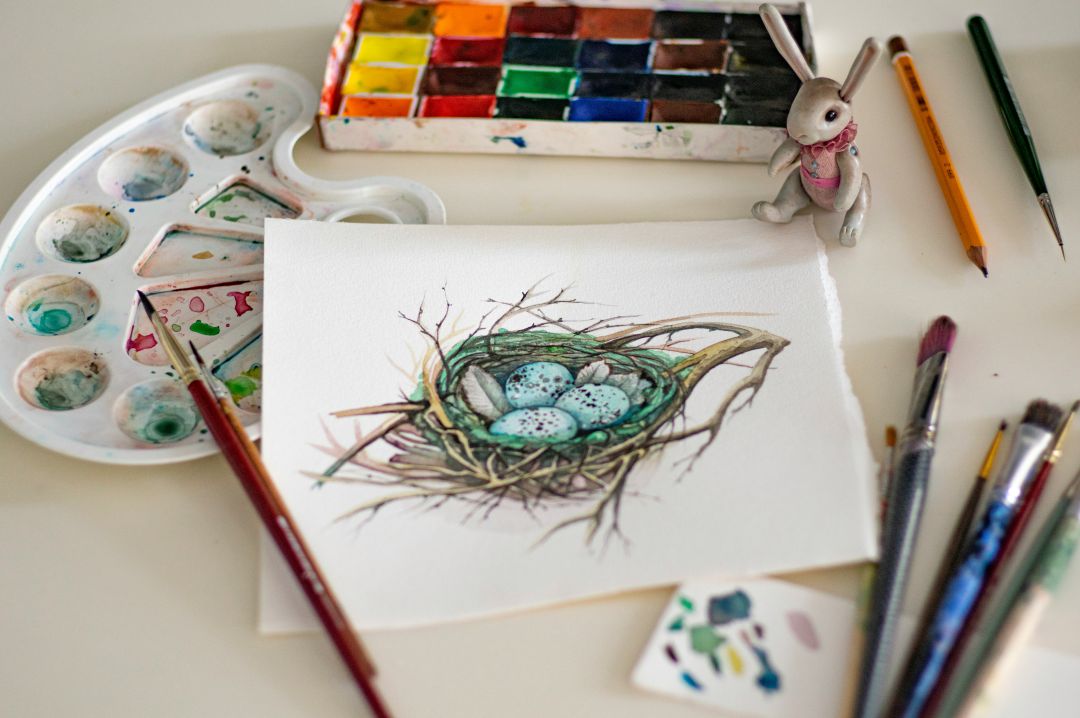 An artists set up, paints surrounding a sketch book with a painted image of a birds nest with eggs inside