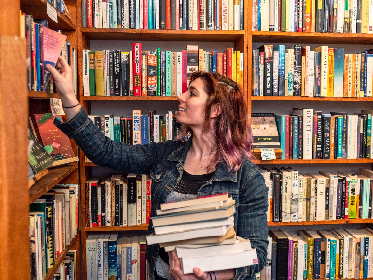 A young woman in the library, one hand holds a stack of books, the other pulling a book from the shelof. The woman looks happy